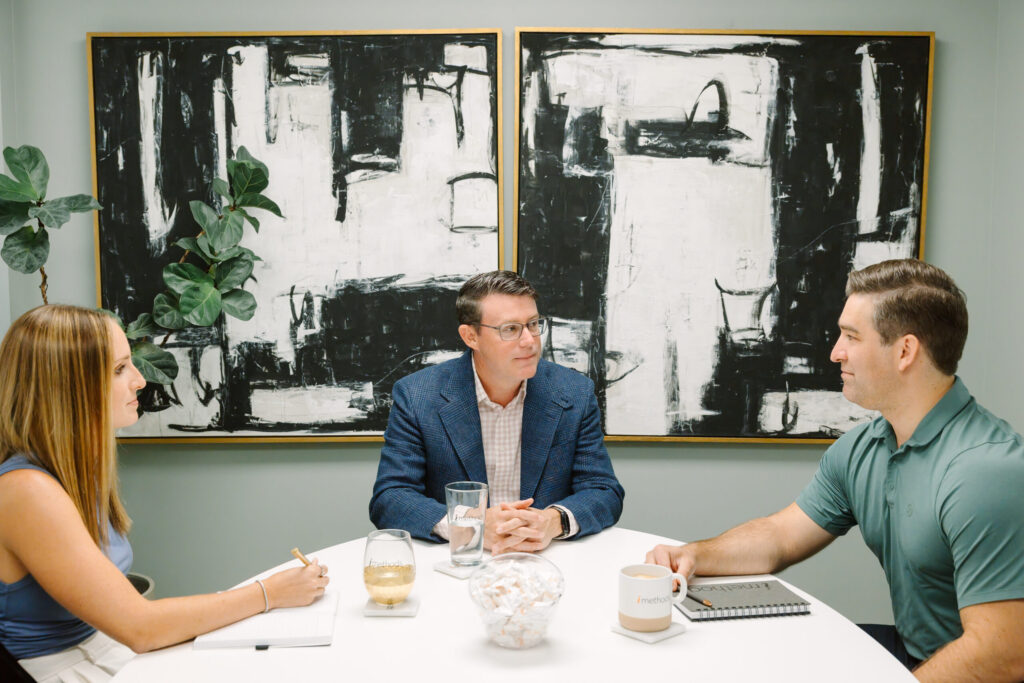 Three people sit around a table in an office, engaged in discussion, with abstract black-and-white artwork and a plant in the background.